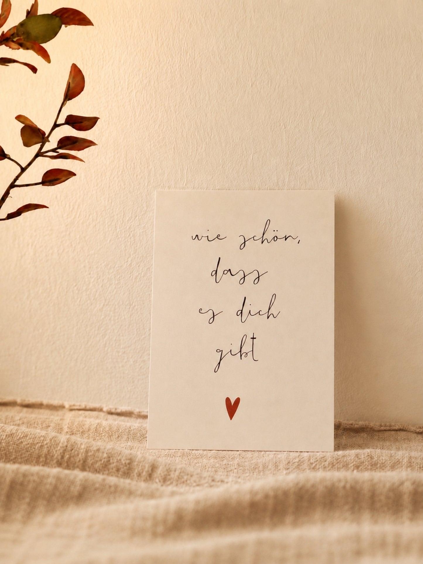 Postkarte "Wie schön, dass es dich gibt" vor warmer Wand mit naturfarbenem Stoff