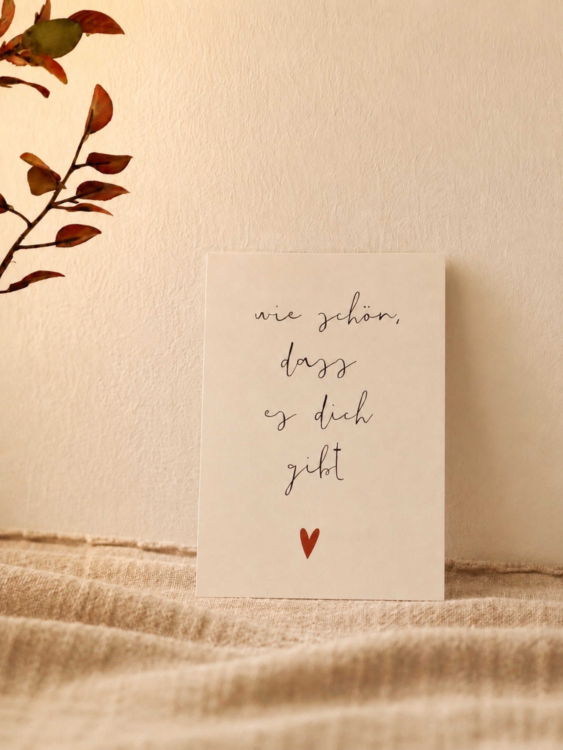 Postkarte "Wie schön, dass es dich gibt" vor warmer Wand mit naturfarbenem Stoff